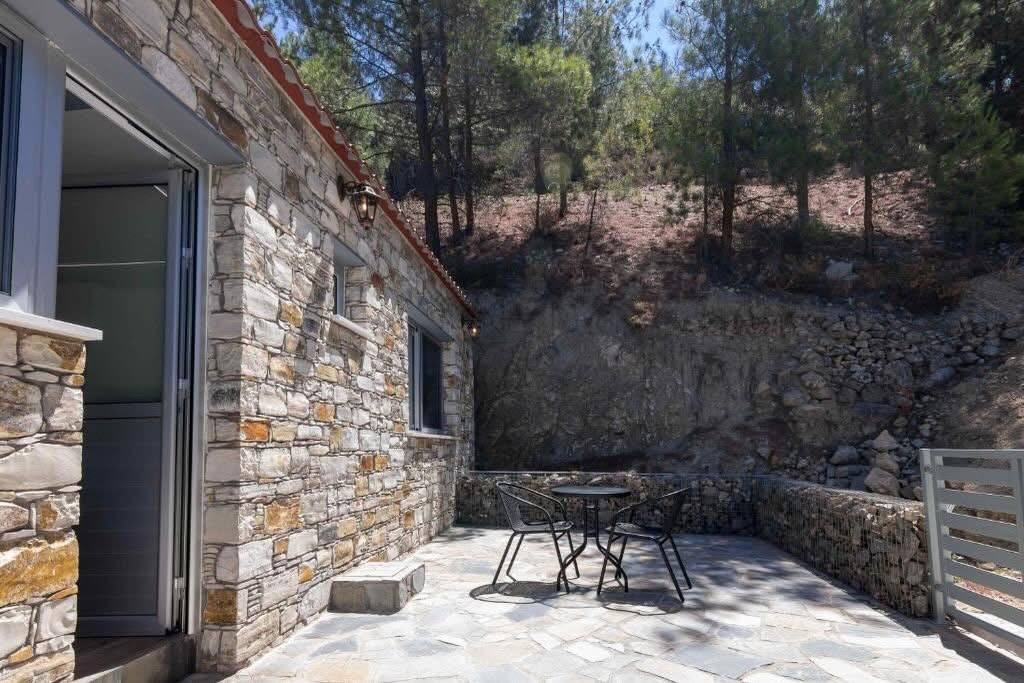 Σκιερή πλαϊνή βεράντα με τραπεζάκι δίπλα στην πέτρινη πρόσοψη και το βραχώδες πρανές